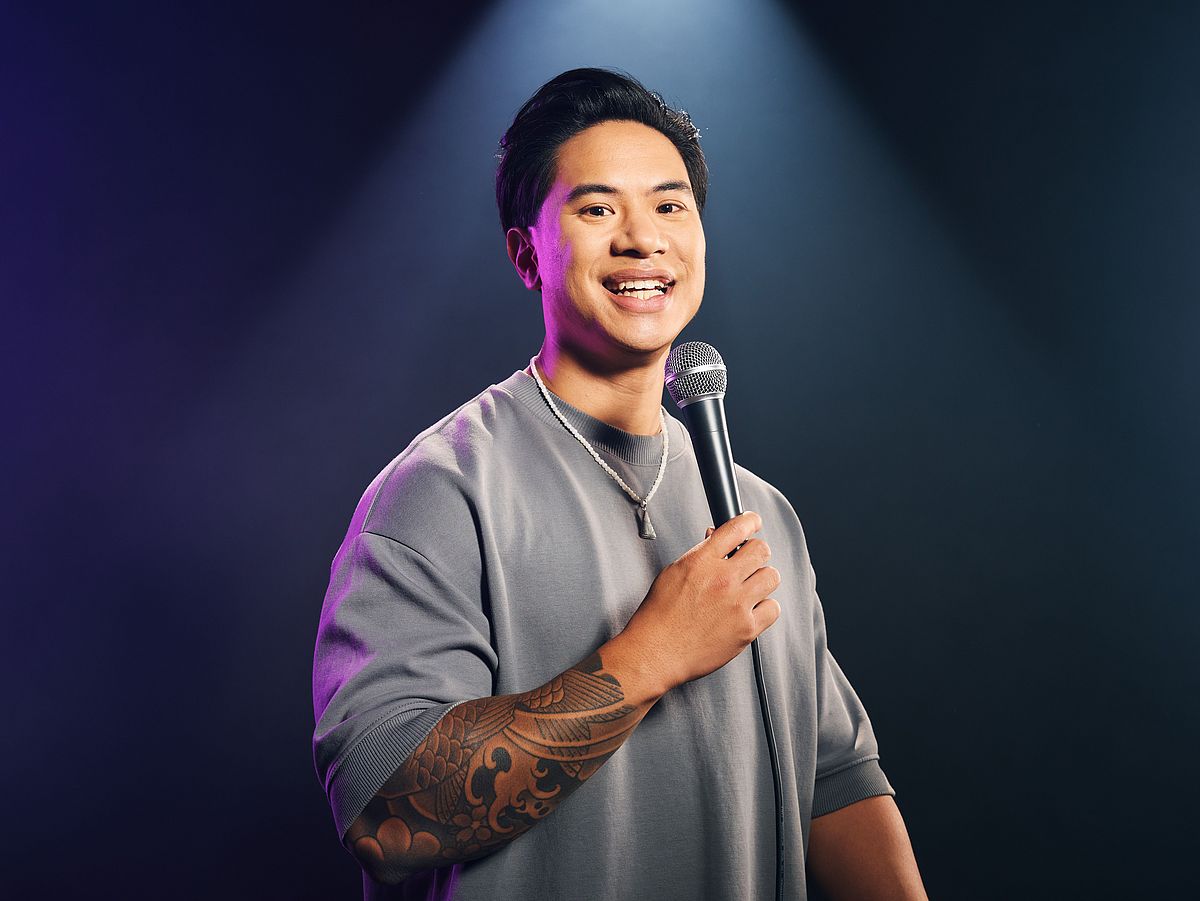 Tutty Tran steht vor einem dunklen Hintergrund. Er trägt einen grauen Pullover. ER hält ein Mikrofon in der Hand. 