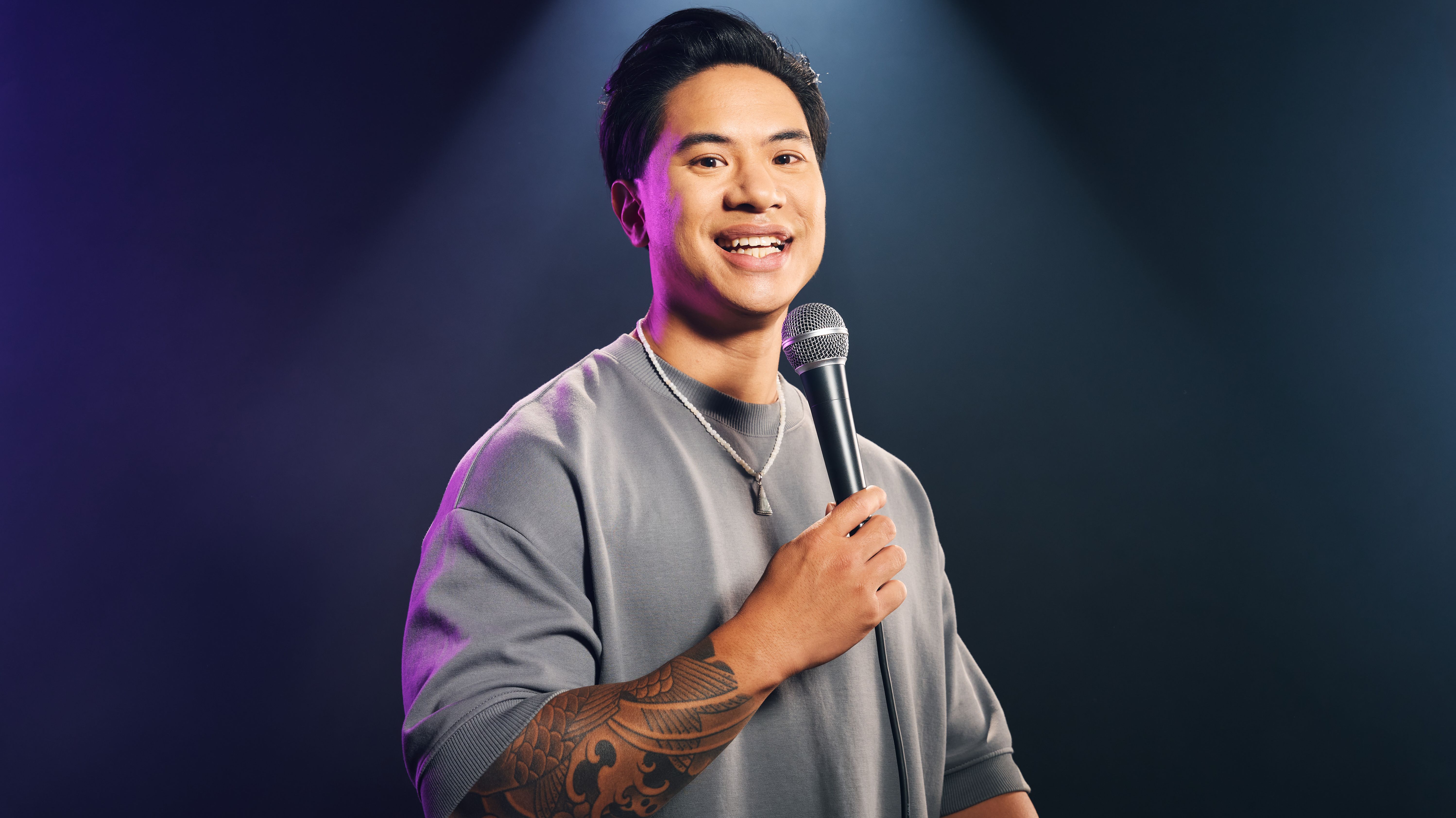 Tutty Tran steht vor einem dunklen Hintergrund. Er trägt einen grauen Pullover. ER hält ein Mikrofon in der Hand. 