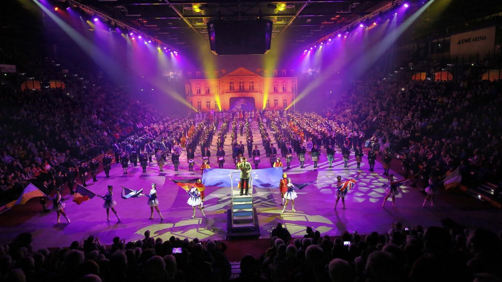 Man blickt auf eine gefüllte Halle, auf einem Podest steht ein Dirigent, auf der Bühnenfläche stehen Musikkapellen. 