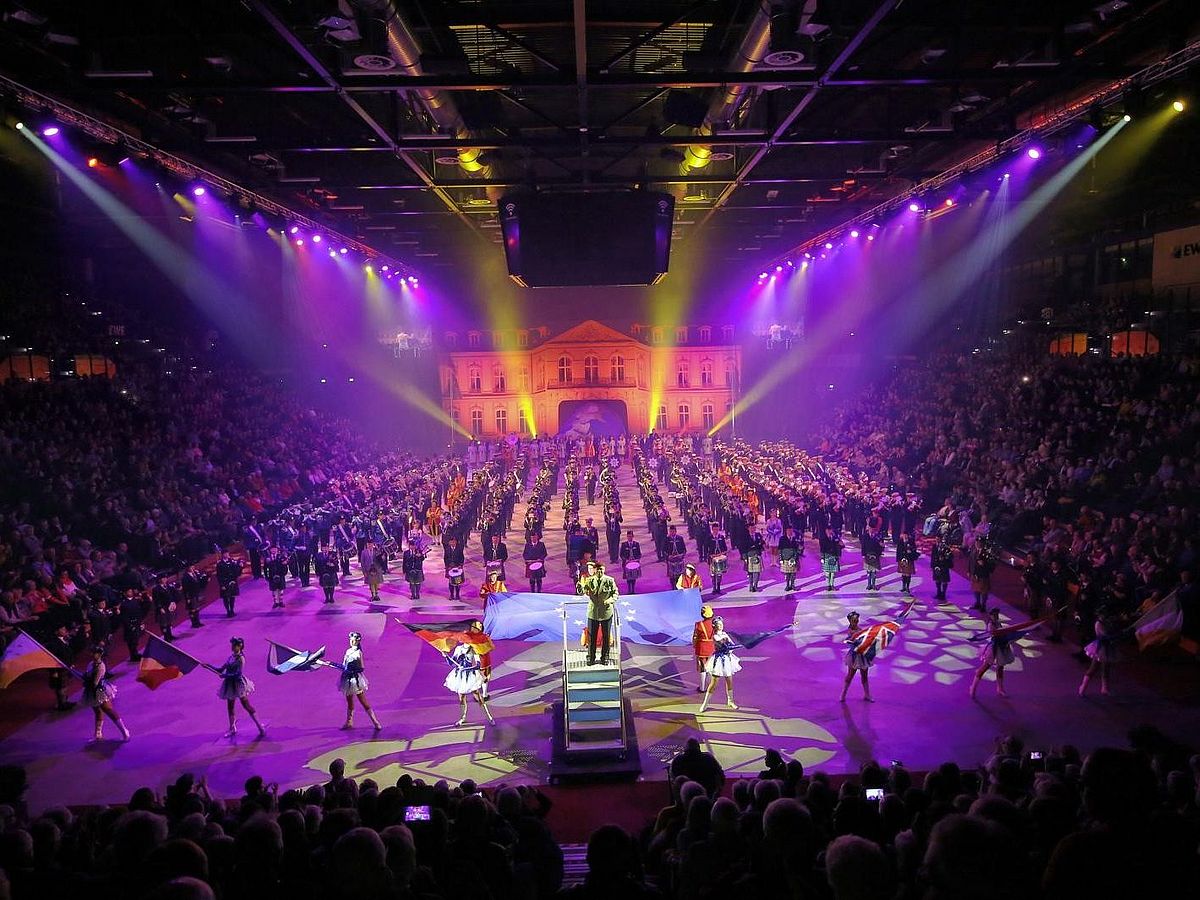 Man blickt auf eine gefüllte Halle, auf einem Podest steht ein Dirigent, auf der Bühnenfläche stehen Musikkapellen. 