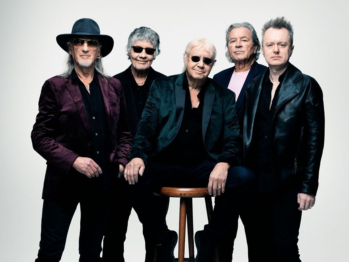 Bandfoto von Deep Purple vor einem weißen Hintergrund. Alle tragen dunkle Kleidung, drei von ihnen haben eine Sonnenbrille an. Einer von ihnen sitzt auf einem Barhocker. 