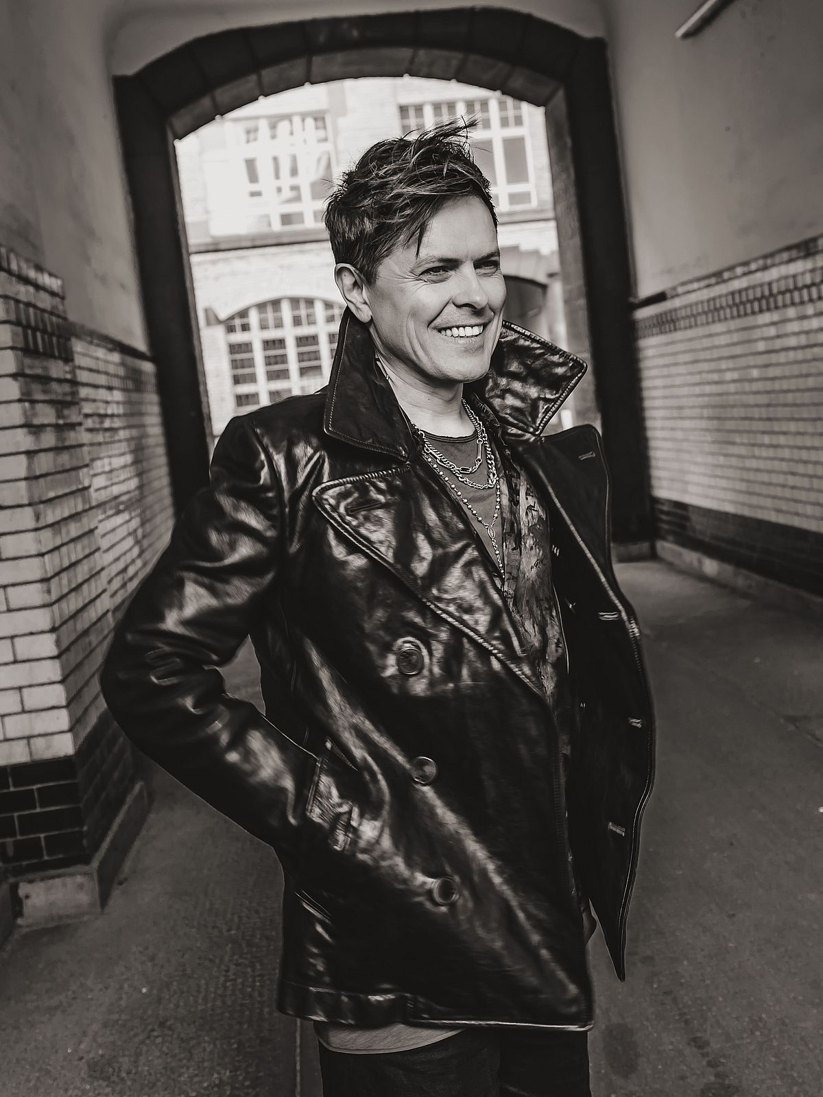 Michael P. Kelly hat eine Lederjacke an. Er steht in einer Hofeinfahrt. 