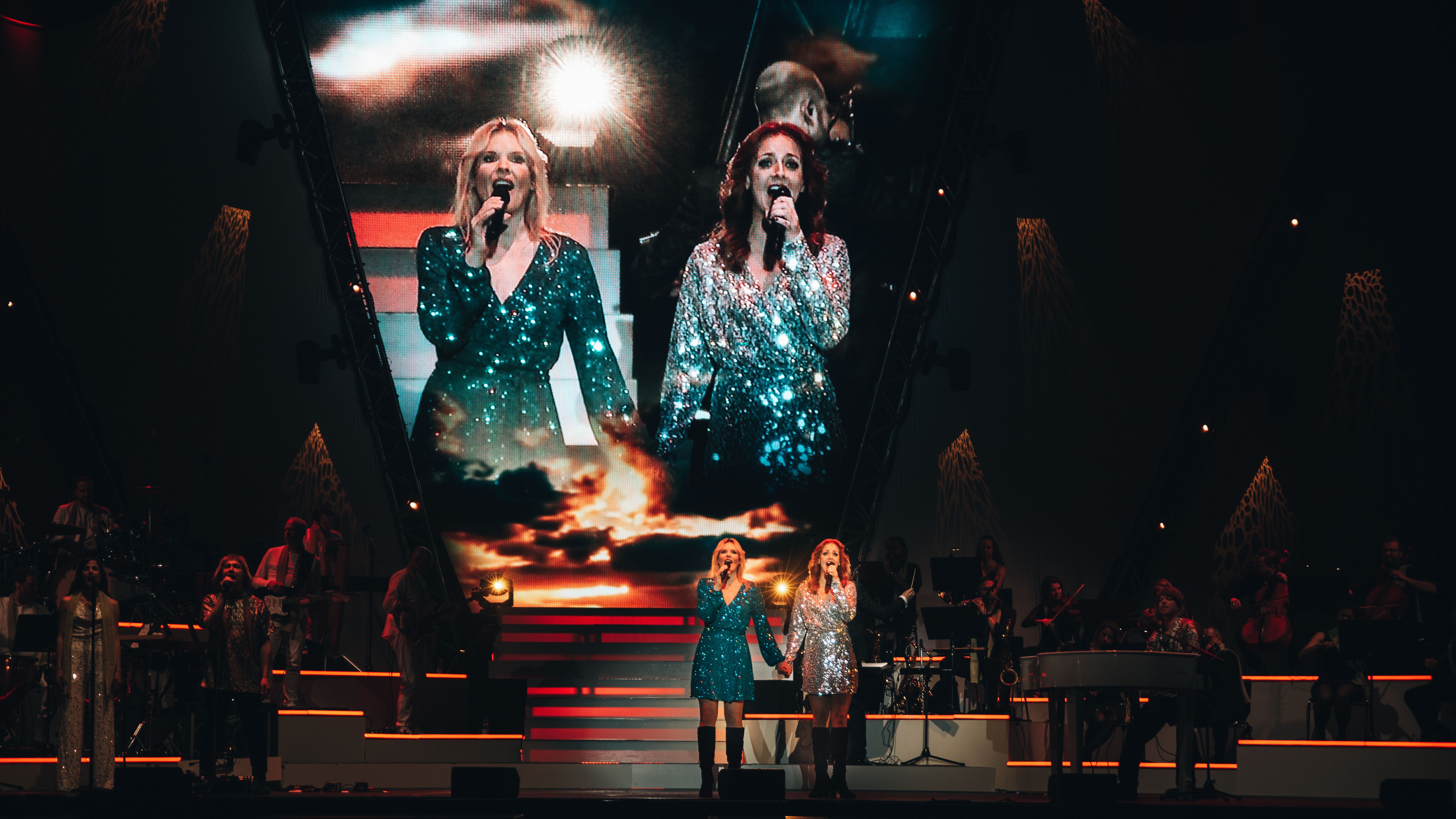 Zwei Sängerinnen stehen auf der Bühne und halten sich an der Hand. Sie tragen beide kurze glitzernde Kleider. 