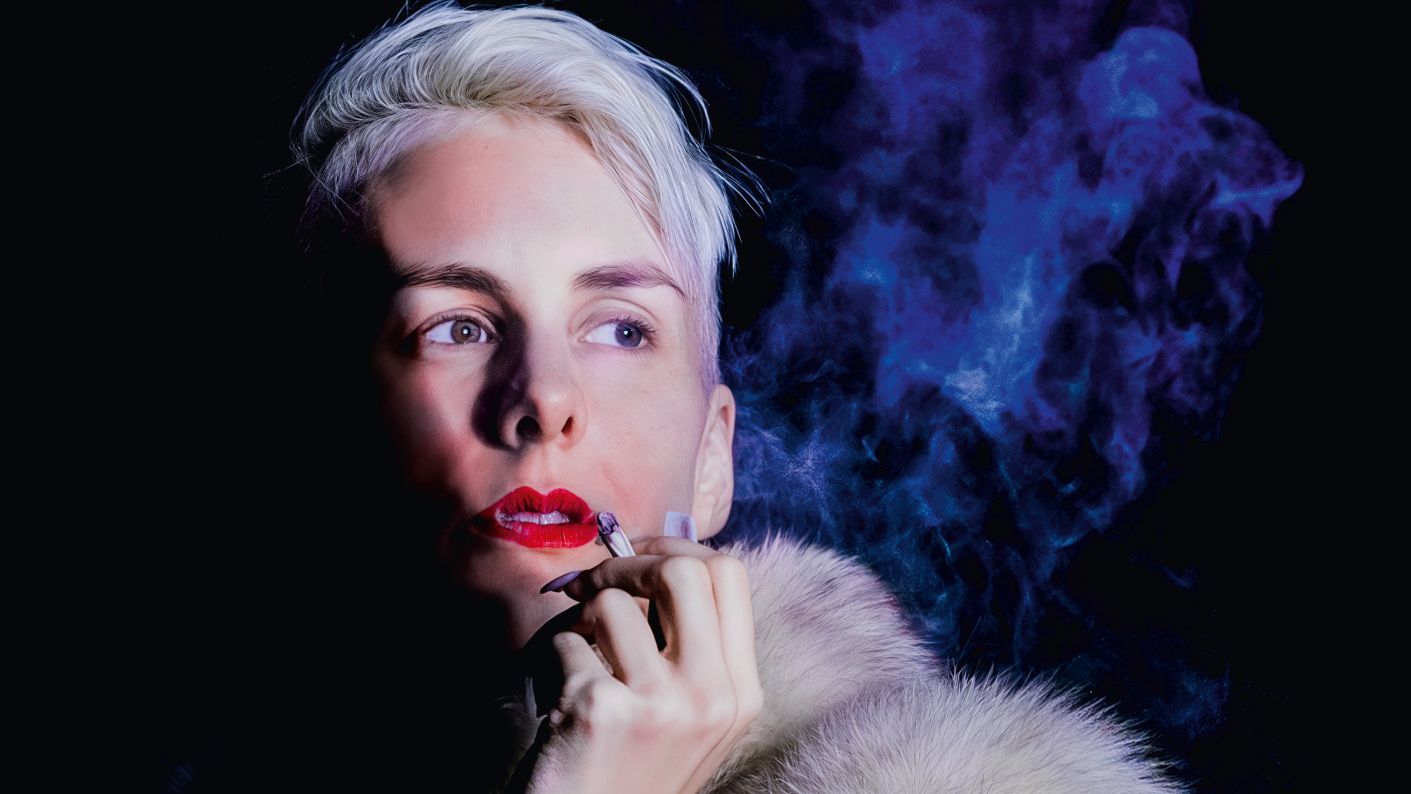 Lisa Eckhart mit rotem Lippenstift trägt einen weißen Pelz und hält eine Zigarette in der Hand.