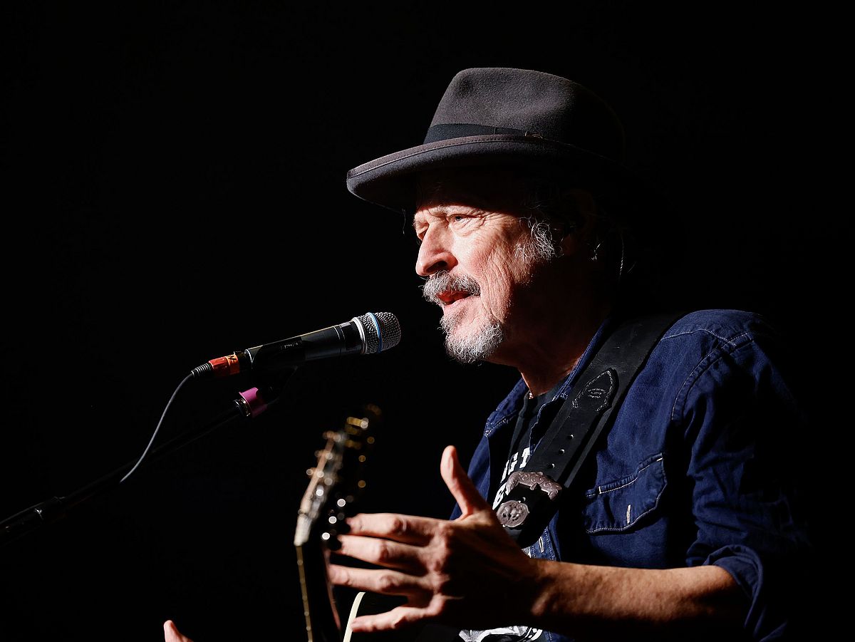 Wolfgang Niedecken steht mit einer Gitarre auf der Bühne. Vor ihm ist ein Mikrofon. Er trägt einen Hut. 