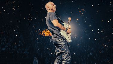 Eros Ramazotti steht mit einer E-Gitarre auf einer Bühne. Er trägt ein T-Shirt und eine Lederhose. Vor ihm ist das Publikum zu sehen, die mit ihren Handys leuchten. 