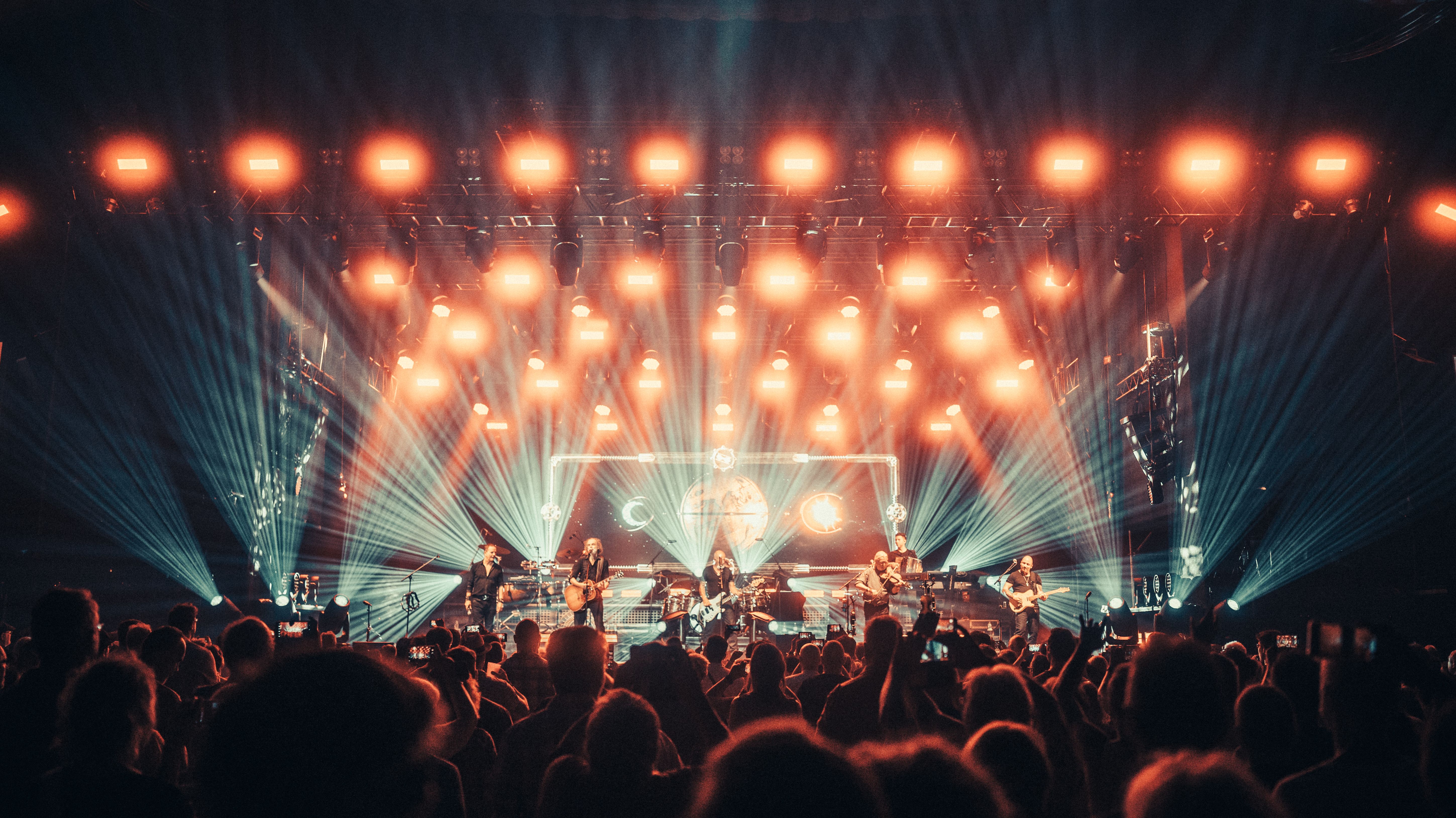 Ein Live-Bild von einem Konzert. Im Vordergrund sind Konzertgäste zu sehen, auf der Bühne steht die Band. Das Licht ist rötlich. 