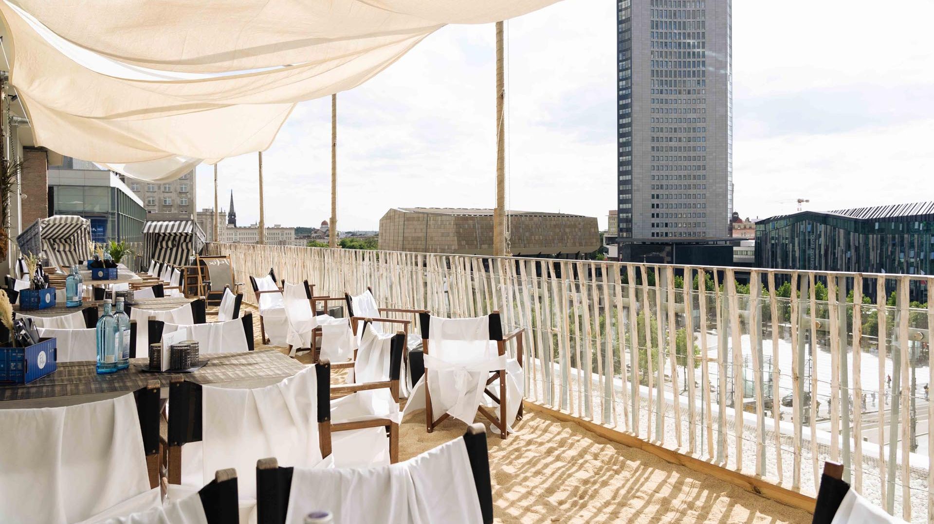Blick von der Hotelterasse mit gedeckten Tischen und Strandkörben auf Sand. Man blickt auf den MDR Tower und auf das Gewandhaus. 