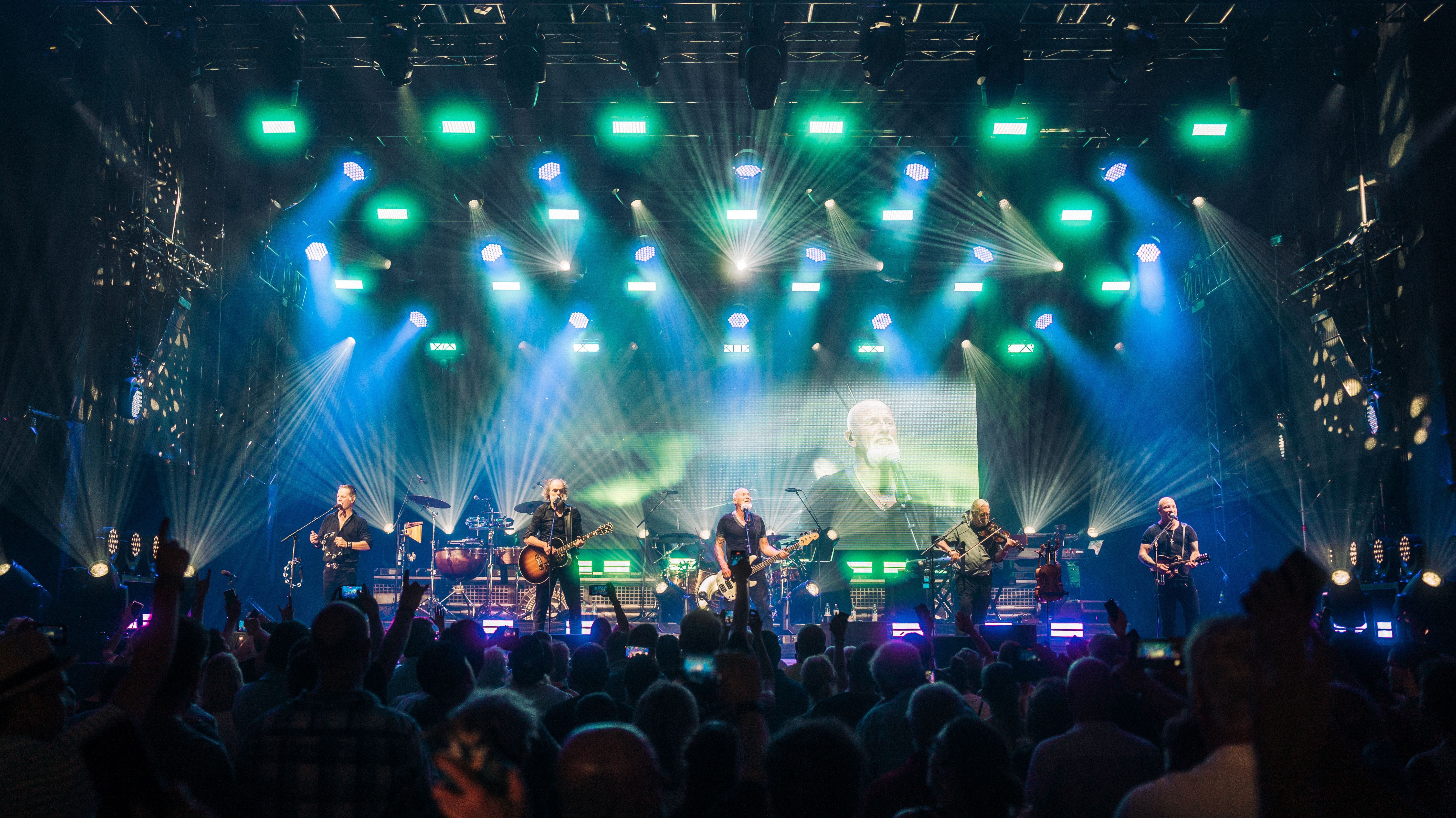 Ein Live-Bild von einem Konzert. Im Vordergrund sind Konzertgäste zu sehen, auf der Bühne steht die Band. Das Licht ist bläulich.