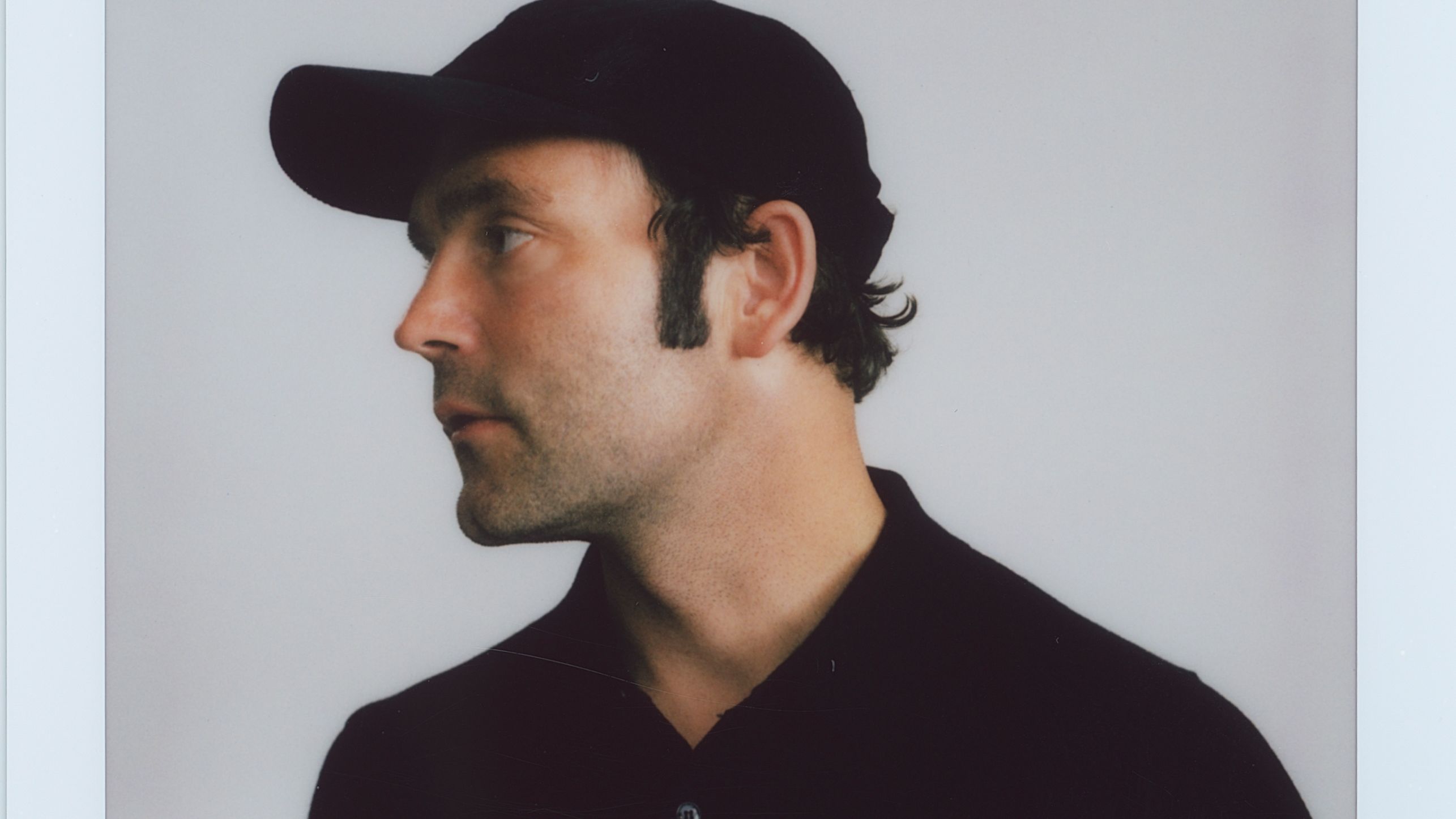 Ein Polaroidfoto von Bosse, der ein schwarzes Käppi und ein schwarzes T-Shirt trägt. Er steht vor einem weißen Hintergrund und schaut nach links. 