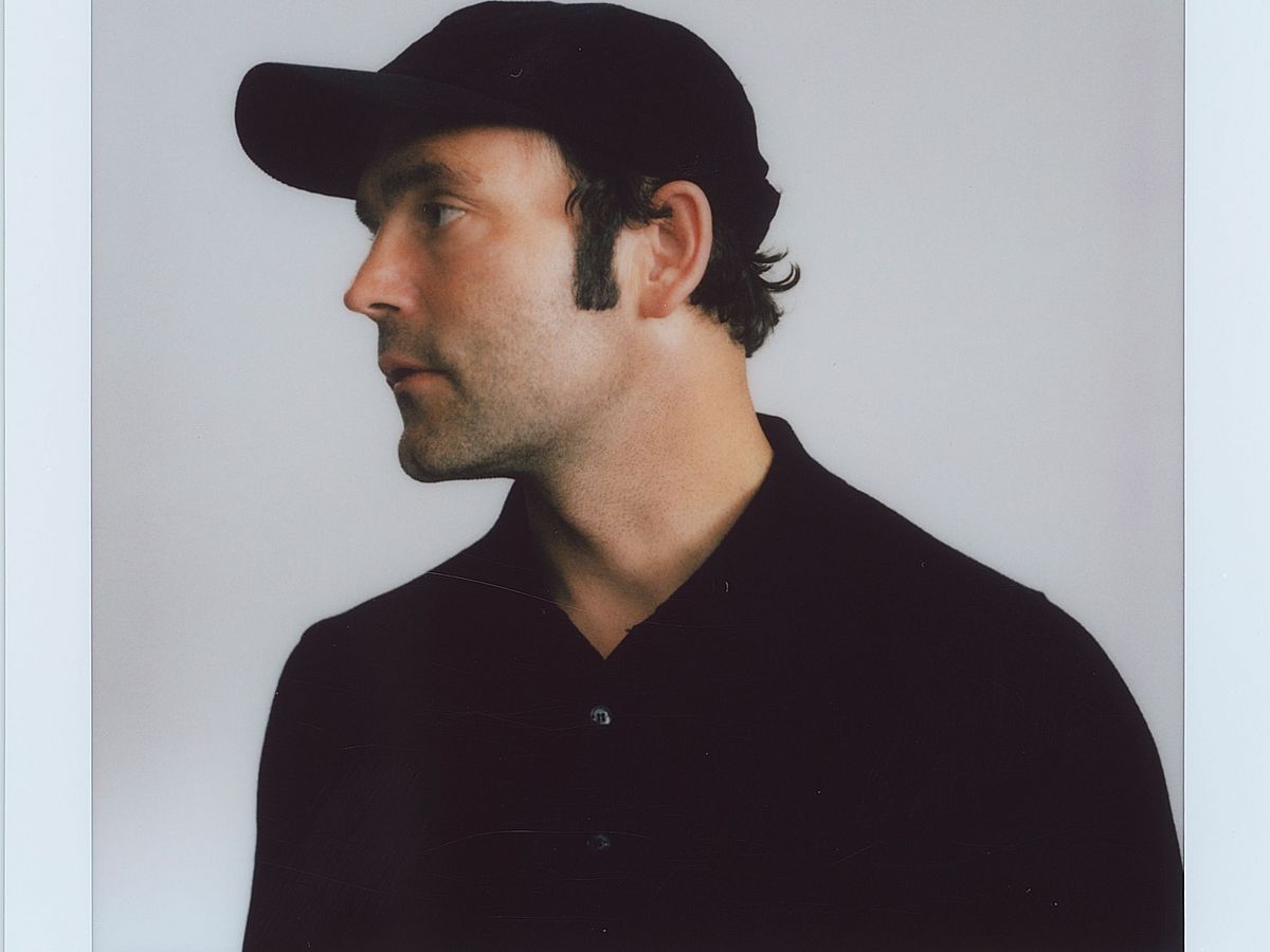 Ein Polaroidfoto von Bosse, der ein schwarzes Käppi und ein schwarzes T-Shirt trägt. Er steht vor einem weißen Hintergrund und schaut nach links. 