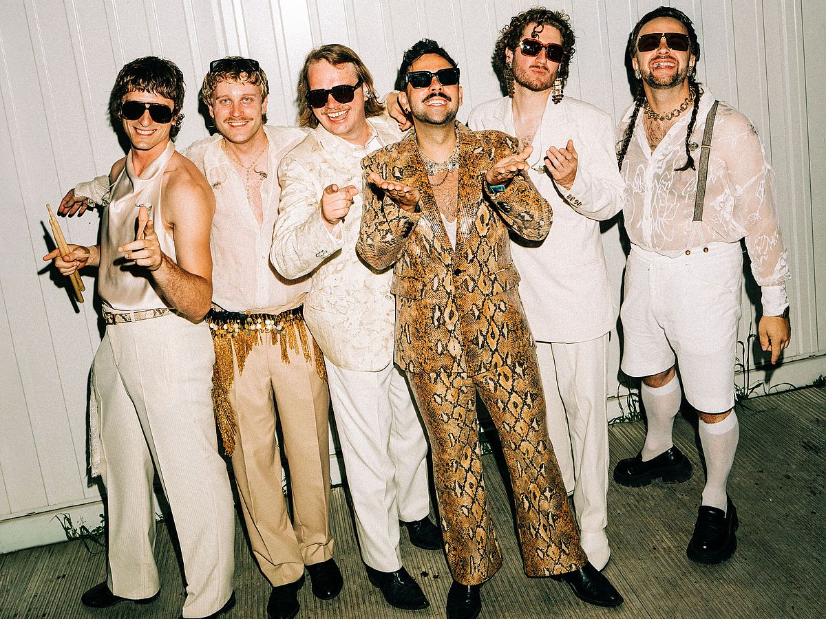 Sechs Männer posieren nebeneinander vor einer hellen Wand. Sie tragen auffällige Sonnenbrillen und extravagante Outfits in Weiß, Beige und Braun. Ein Mann in der Mitte sticht mit einem Anzug im Schlangenhautmuster hervor. Die Gruppe lächelt und gestikuliert lässig in die Kamera.