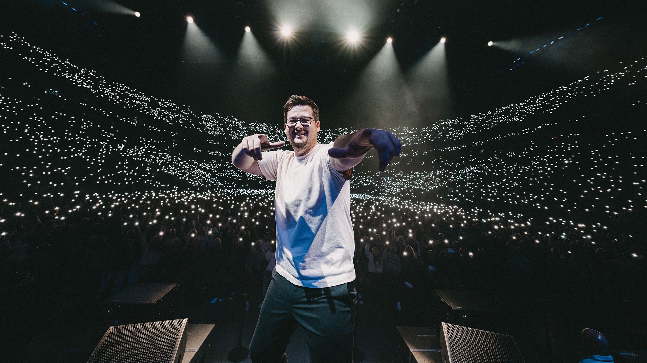 Chris Tall steht lächelnd auf der Bühne, im Hintergrund sind in einer große Halle viele Handylichter zu sehen. 