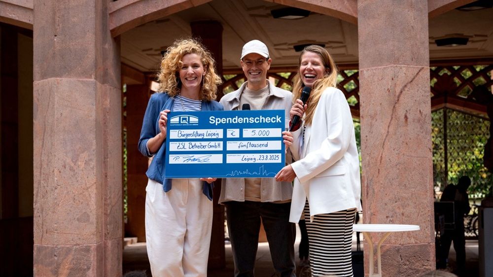 Trendela Braun, Philipp Franke und die Moderatorin stehen im Außenbereich des Grassimuseums und halten einen Spendenscheck hoch. 