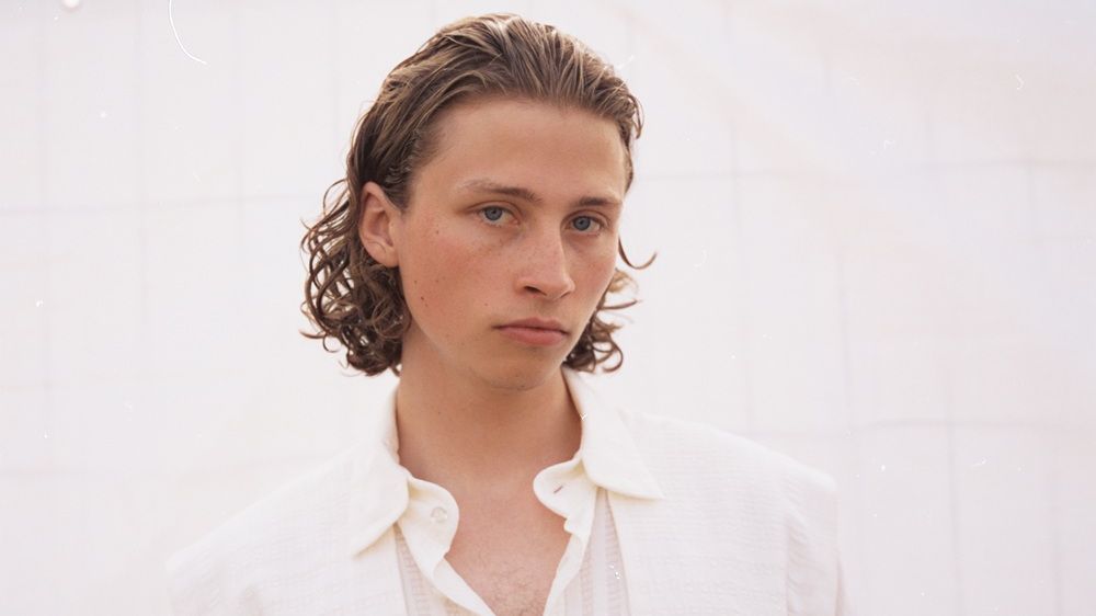 Berq steht vor einer weißen Wand und schaut in die Kamera. Er trägt ein weißes Hemd. 