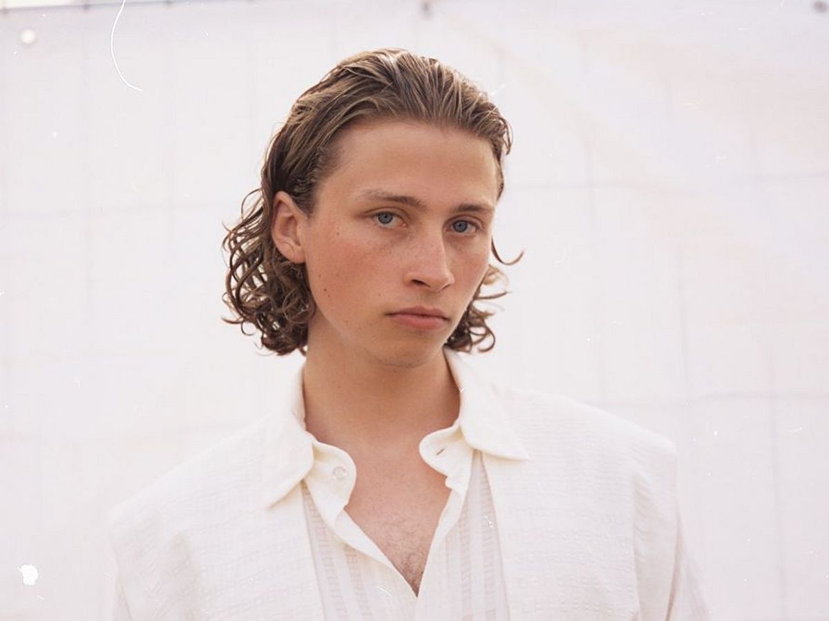 Berq steht vor einer weißen Wand und schaut in die Kamera. Er trägt ein weißes Hemd. 