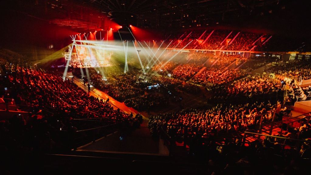 Blick auf eine gefüllte Arena auf eine Bühne mit Lichtershow.