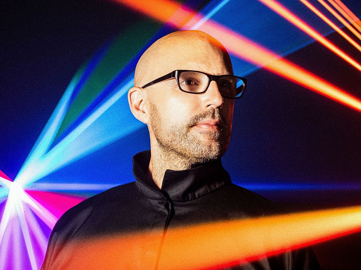 Schiller trägt eine Brille und einen dunklen Pulli, oranges und blaue Strahler sind zu sehen. 