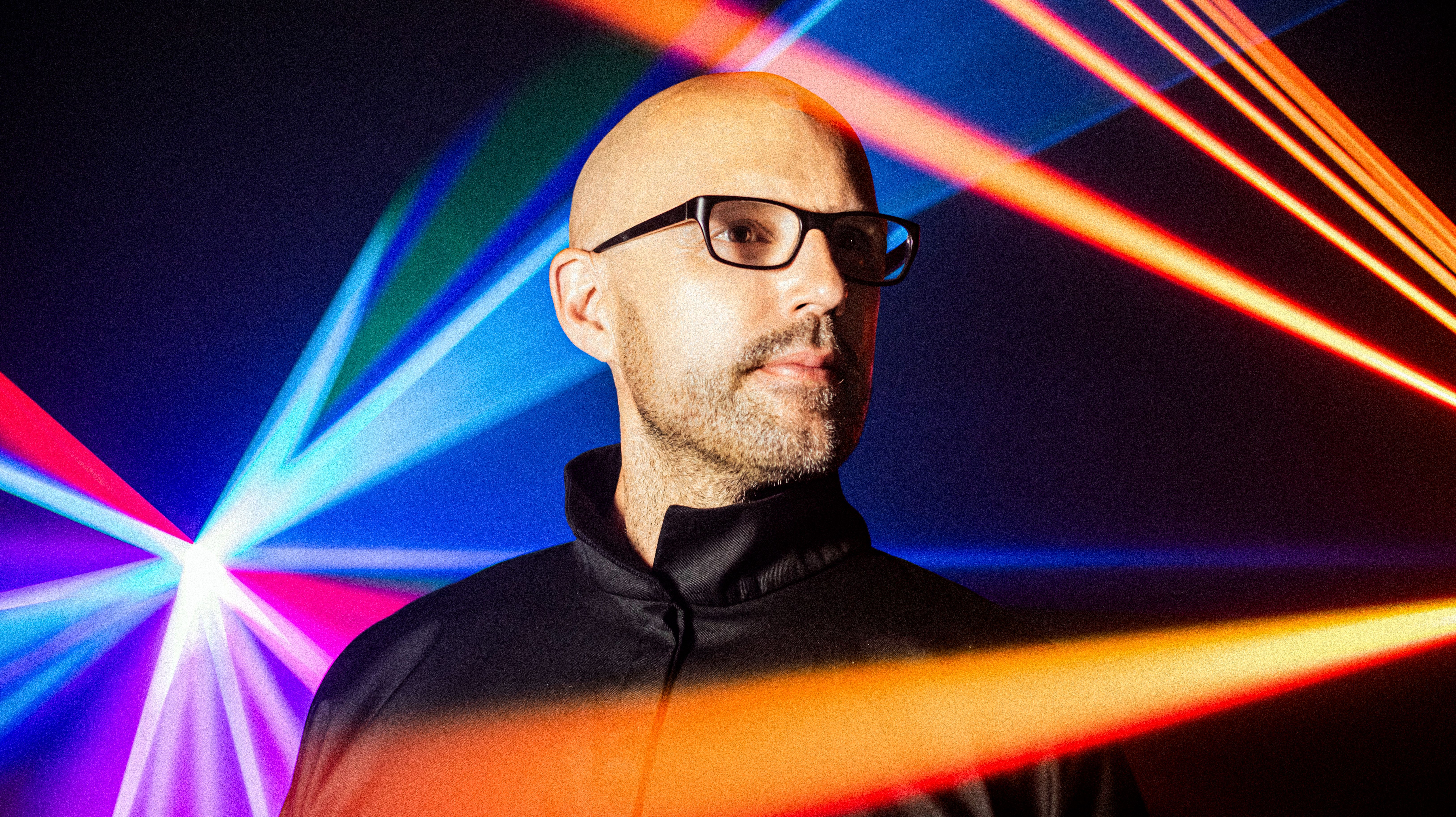 Schiller trägt eine Brille und einen dunklen Pulli, oranges und blaue Strahler sind zu sehen. 