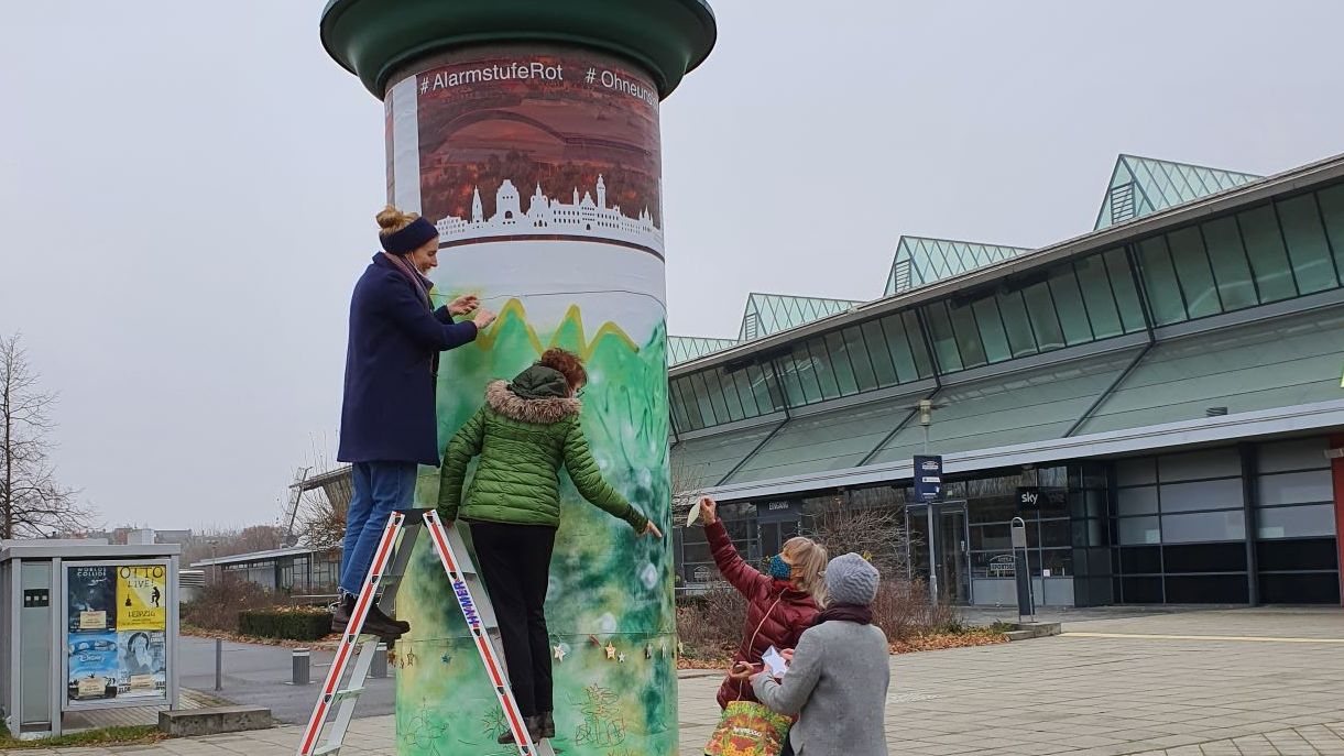 Zwei Frauen auf einer Leiter bringen gebastelte Stern an einer bunten bemalten Litfaßsäule an.