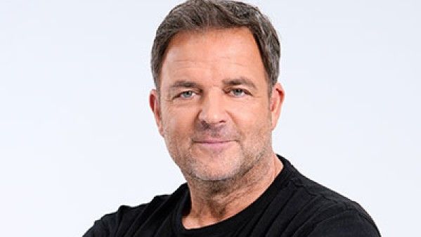 Martin Rütter trägt ein schwarzes T-Shirt, er hat die Arme ineinander verschränkt und lächelt in die Kamera- Er steht vor einem weißen Hintergrund. 