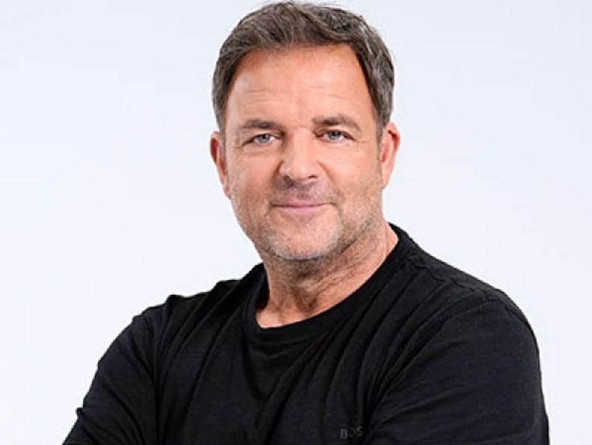 Martin Rütter trägt ein schwarzes T-Shirt, er hat die Arme ineinander verschränkt und lächelt in die Kamera- Er steht vor einem weißen Hintergrund. 