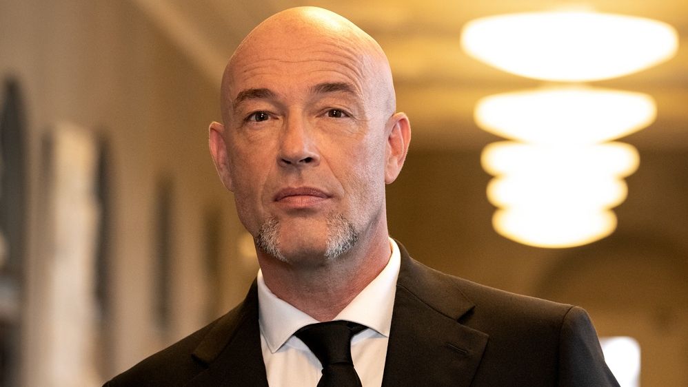 Der Graf steht in einem schwarzen Anzug in einem beleuchtenden Raum.