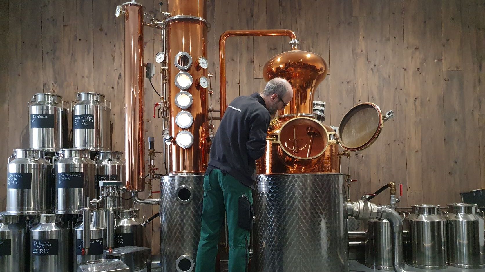 Ein Mann vor einer Destillationsanlage in einem Raum mit mehreren Metallfässern.