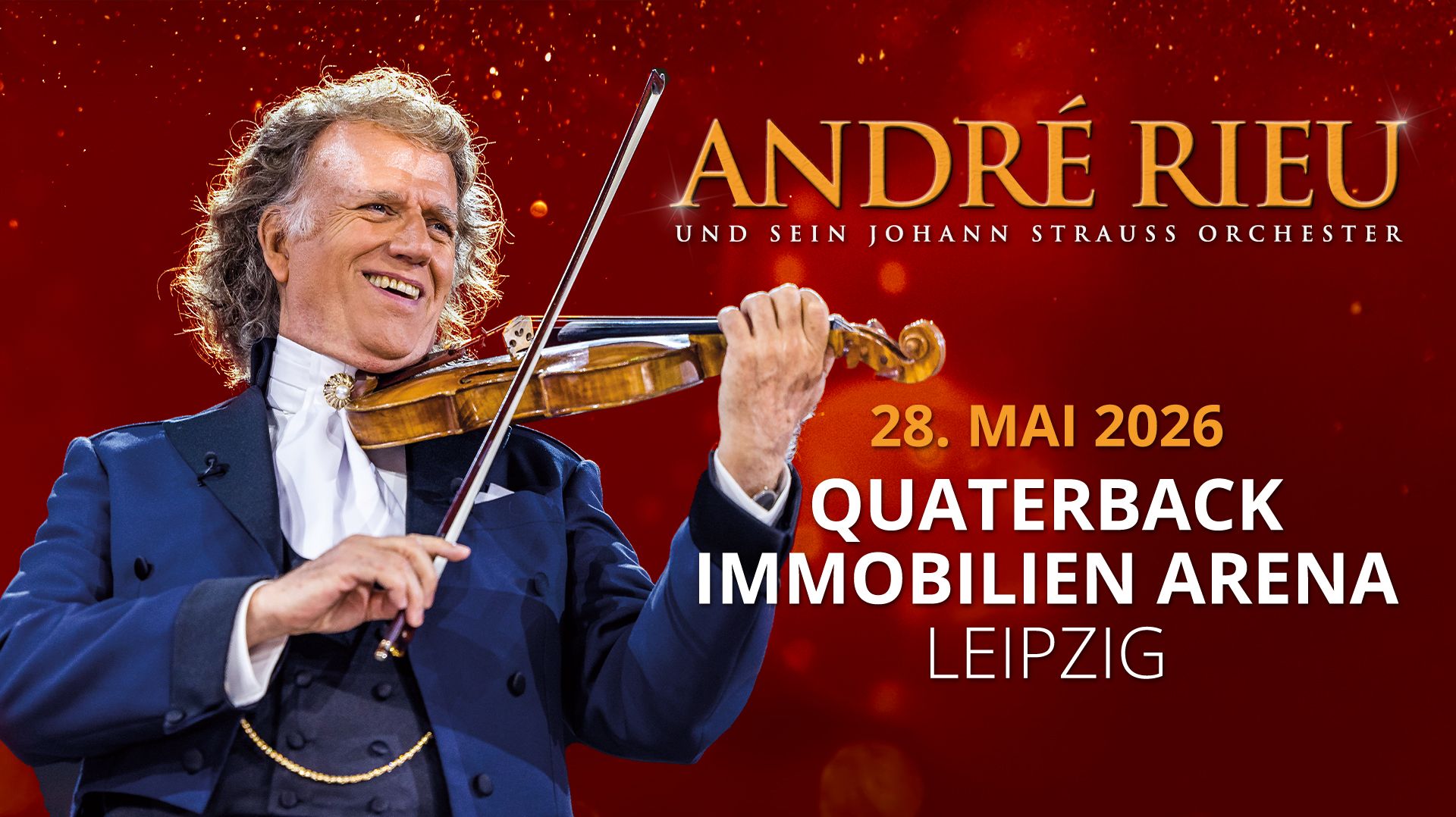 André Rieu trägt einen blauen Anzug mit einem weißen Hemd. Er hält eine Geige. 