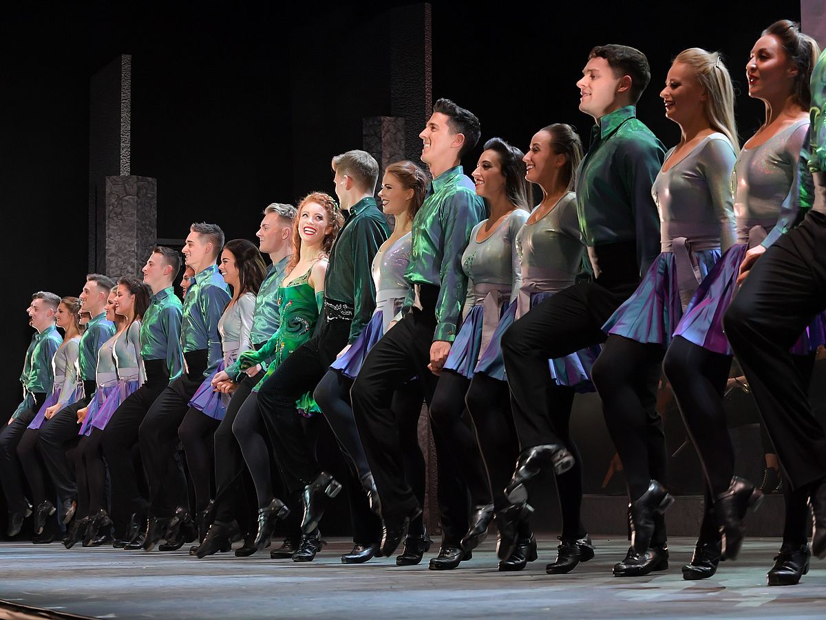 Die Tänzerinnen und Tänzer stehen in einer langen Reihe. Sie stehen alle auf einem Bein, das zweite sind sie gerade dabei, es nach oben zu heben. Alle lächeln. Die Männer tragen grüne, glänzende Hemden und schwarze Hosen, die Frauen tragen lila Röcke und ein helles Oberteil. 