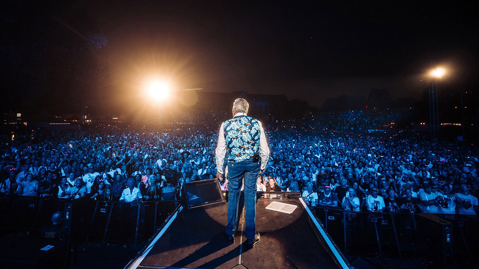 Es ist ein Live-Bild eines Open-Air-Konzertes von Roland Kaiser. Das Bild wurde von der Bühne aus aufgenommen, es ist dunkel mit zwei hellen Strahlern auf Roland Kaiser. Er steht in der Mitte der Bühne und trägt eine bunte Weste mit weißen Hemd. Vor ihm jubelt das Publikum. 