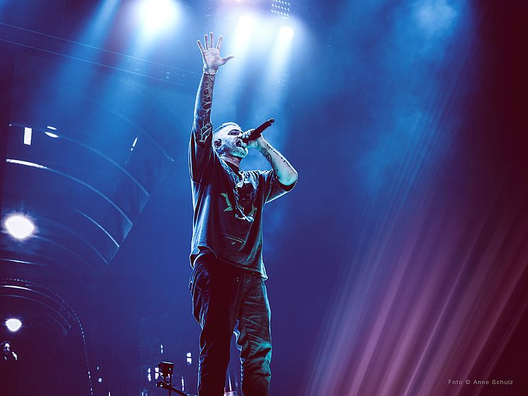 Bushido hat ein Mikrofon vor seinem Mund und streckt den anderen Arm nach oben. Alles ist in dunkelblau gehalten. 