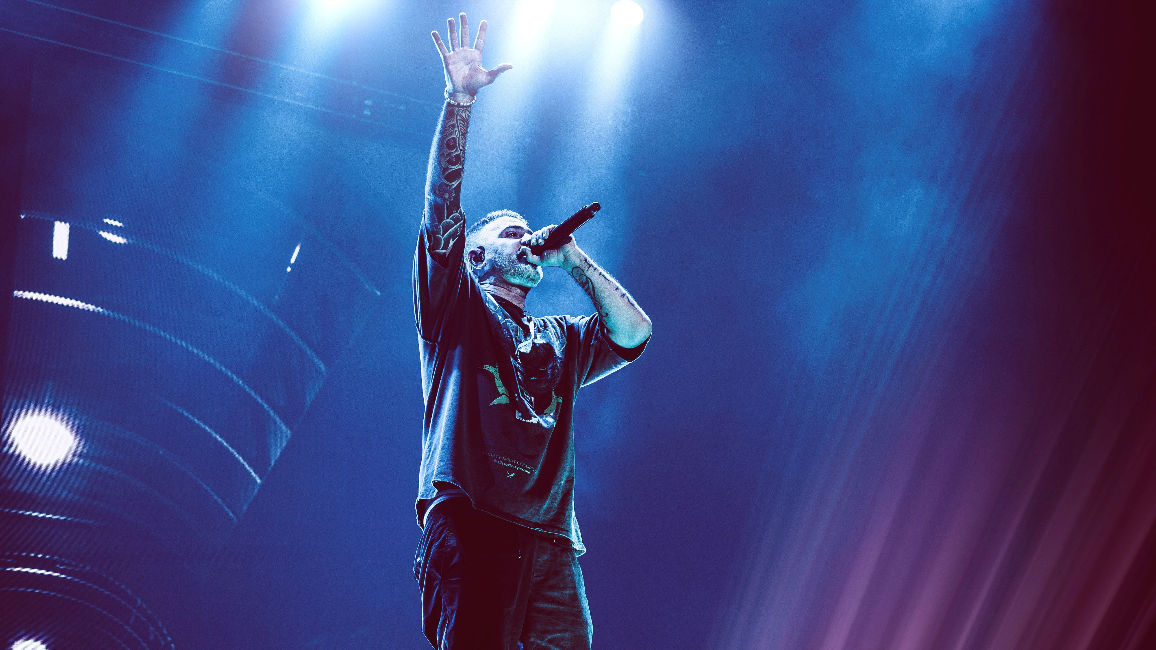 Bushido hat ein Mikrofon vor seinem Mund und streckt den anderen Arm nach oben. Alles ist in dunkelblau gehalten. 