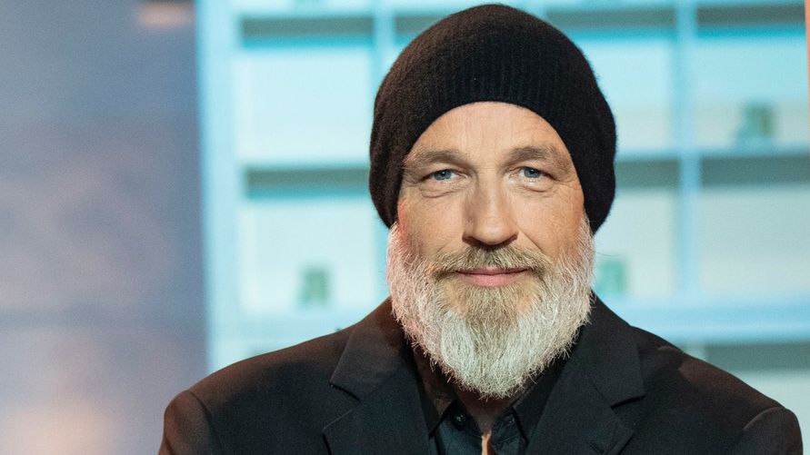 Torsten Sträter hat einen Bart und trägt eine schwarze Mütze sowie einen schwarzen Anzug. Er steht vor einem bläulichem Regal, das im Hintergrund unscharf zu sehen ist. 