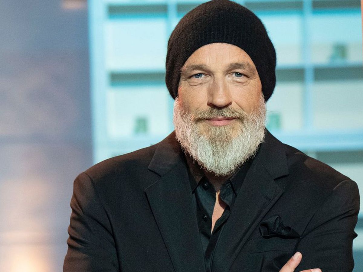 Torsten Sträter hat einen Bart und trägt eine schwarze Mütze sowie einen schwarzen Anzug. Er steht vor einem bläulichem Regal, das im Hintergrund unscharf zu sehen ist. 