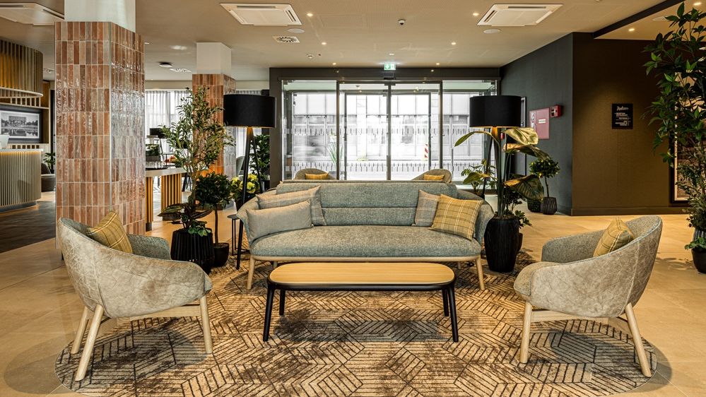 Blick in den Lobbybereich des Hotels. Im Vordergrund sind Polstermöbel in grau- und Holzfarben. n 