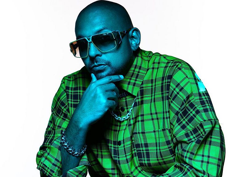 Ein künstlerisch beleuchtetes Porträt eines sitzenden Mannes mit Glatze, großformatiger Sonnenbrille und auffälligem, grün kariertem Hemd. Er stützt sein Kinn nachdenklich mit einer Hand ab, trägt Armbänder und eine Kette, und hat eine entspannte, selbstbewusste Pose. Die blau-grüne Lichtstimmung verleiht dem Bild einen modernen, stylisierten Look.