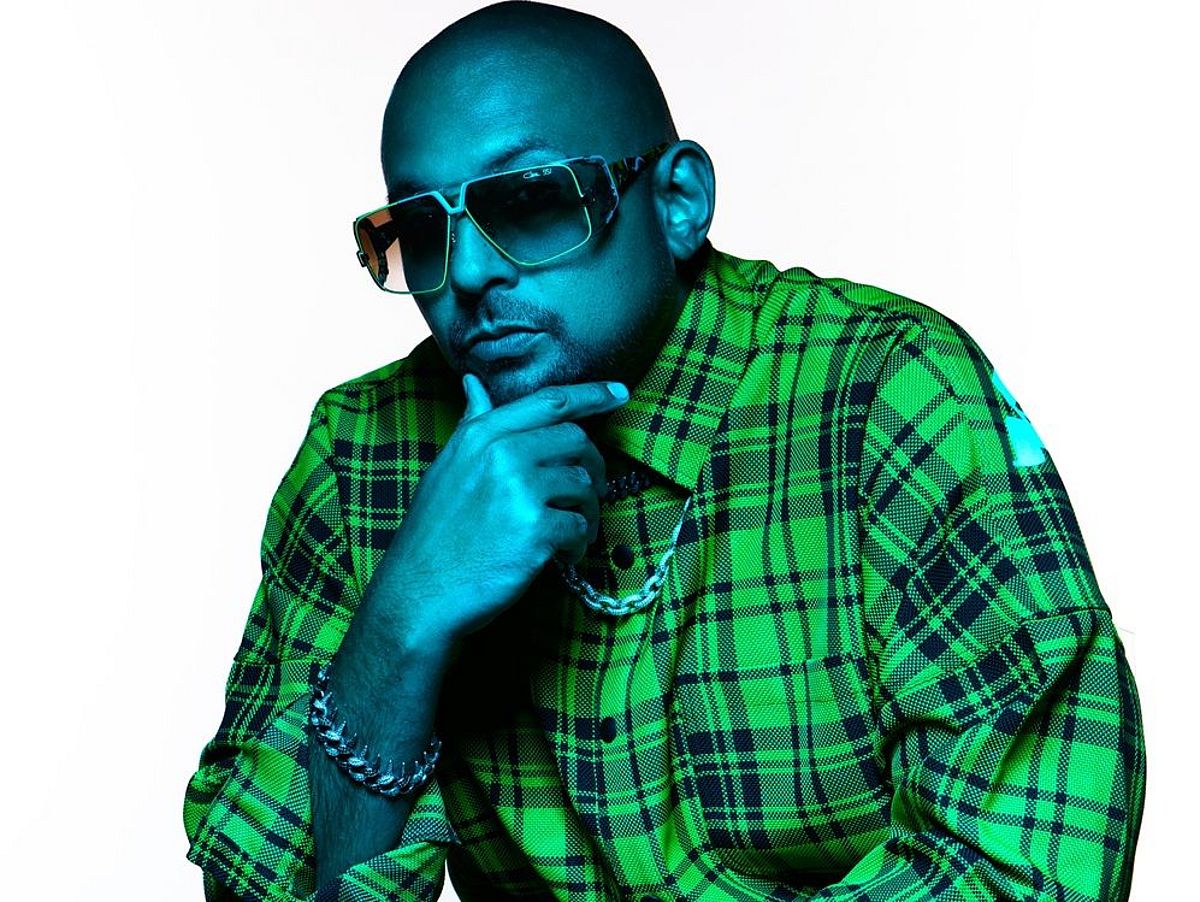 Ein künstlerisch beleuchtetes Porträt eines sitzenden Mannes mit Glatze, großformatiger Sonnenbrille und auffälligem, grün kariertem Hemd. Er stützt sein Kinn nachdenklich mit einer Hand ab, trägt Armbänder und eine Kette, und hat eine entspannte, selbstbewusste Pose. Die blau-grüne Lichtstimmung verleiht dem Bild einen modernen, stylisierten Look.