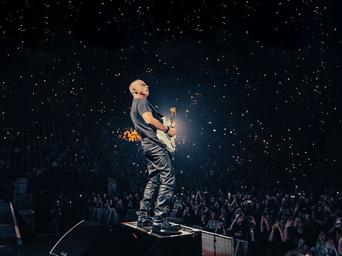 Eros Ramazotti steht mit einer E-Gitarre auf einer Bühne. Er trägt ein T-Shirt und eine Lederhose. Vor ihm ist das Publikum zu sehen, die mit ihren Handys leuchten. 