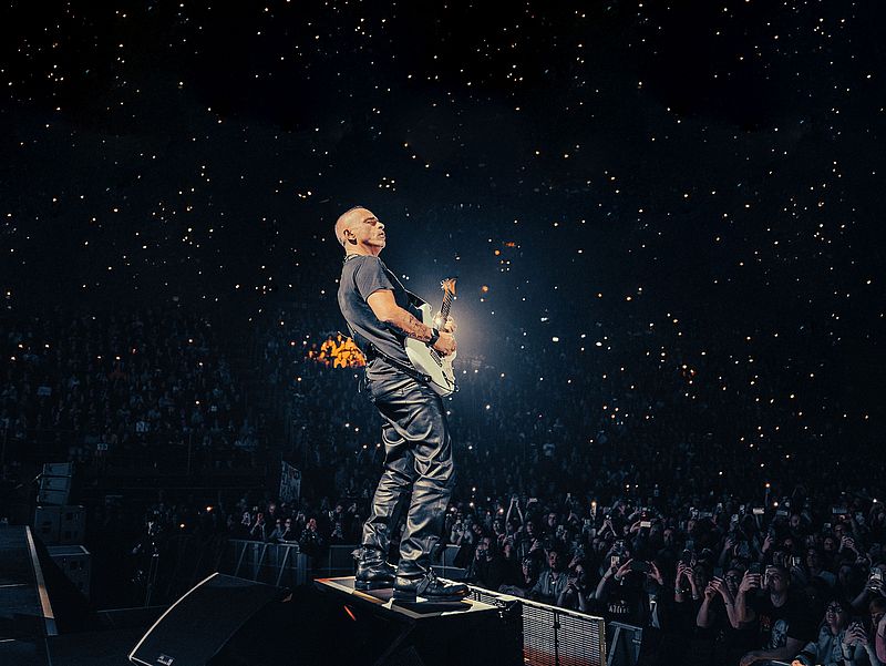 Eros Ramazotti steht mit einer E-Gitarre auf einer Bühne. Er trägt ein T-Shirt und eine Lederhose. Vor ihm ist das Publikum zu sehen, die mit ihren Handys leuchten. 