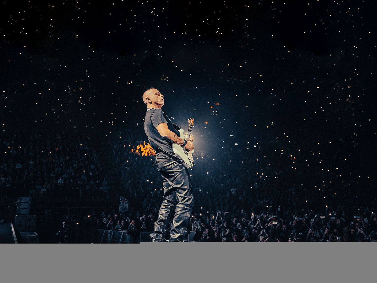 Eros Ramazotti steht mit einer E-Gitarre auf einer Bühne. Er trägt ein T-Shirt und eine Lederhose. Vor ihm ist das Publikum zu sehen, die mit ihren Handys leuchten. 