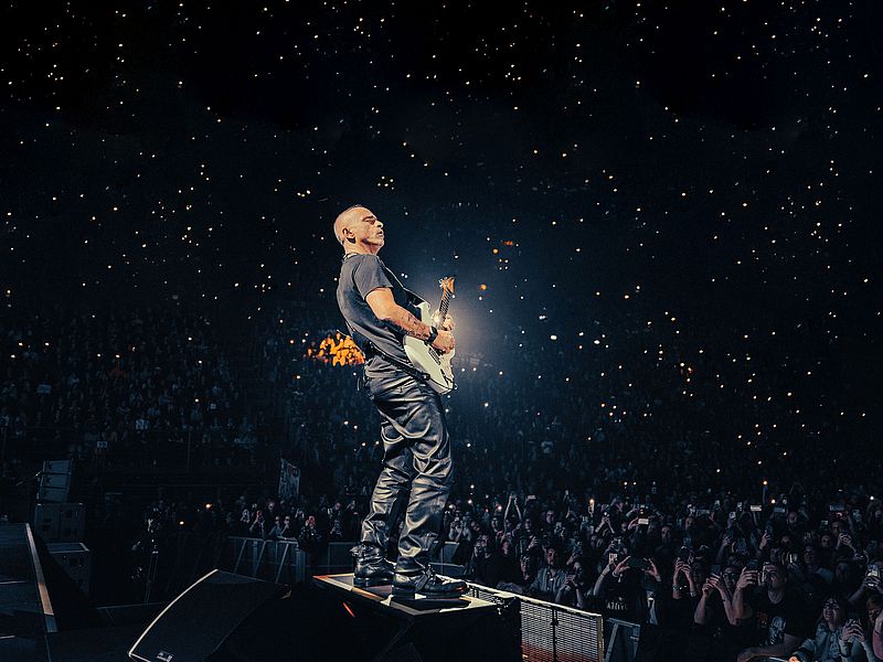 Eros Ramazotti steht mit einer E-Gitarre auf einer Bühne. Er trägt ein T-Shirt und eine Lederhose. Vor ihm ist das Publikum zu sehen, die mit ihren Handys leuchten. 