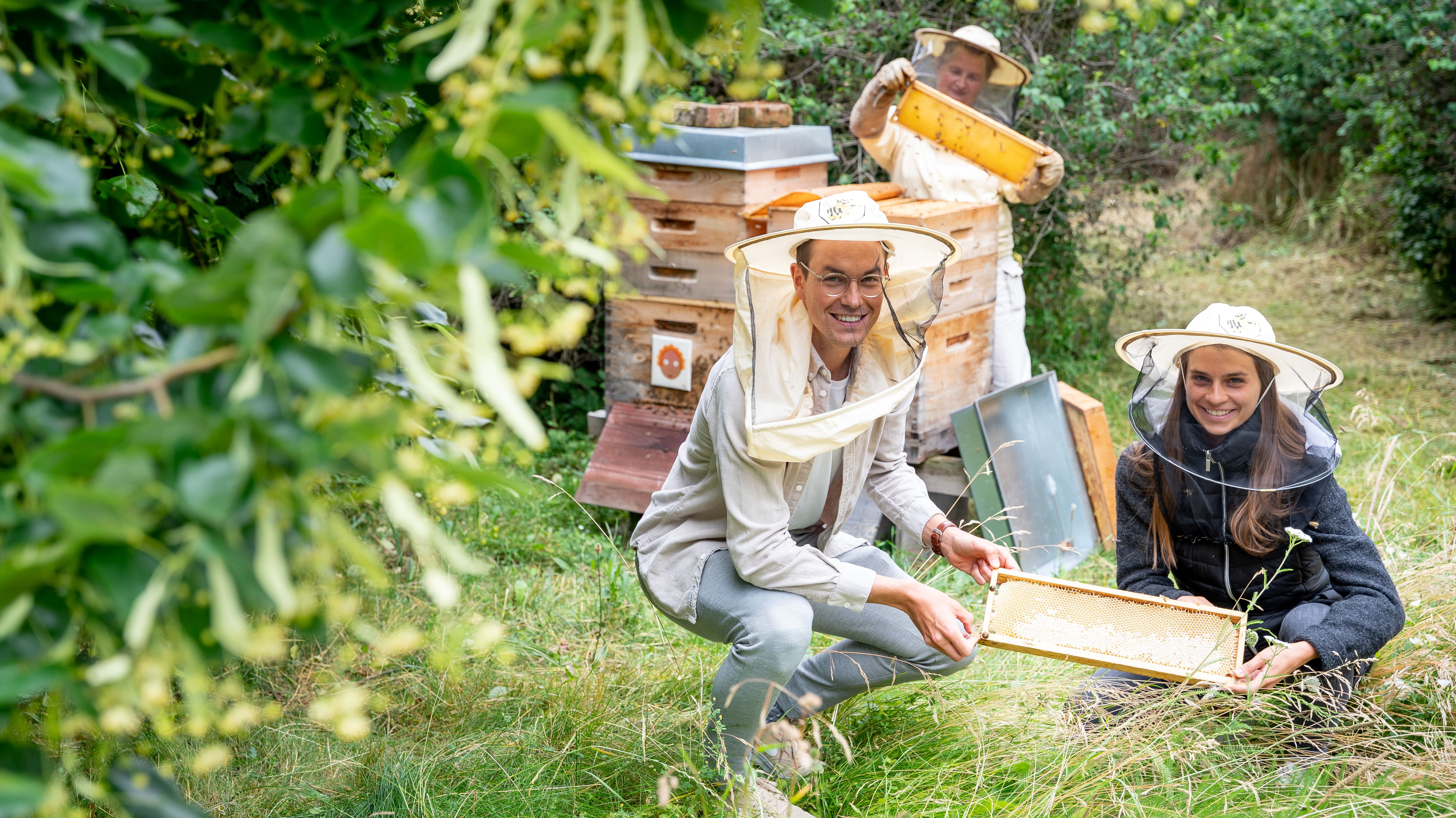 Ein Mann und eine Frau mit Bienenschutzkleidung vor einem Bienenkasten. 