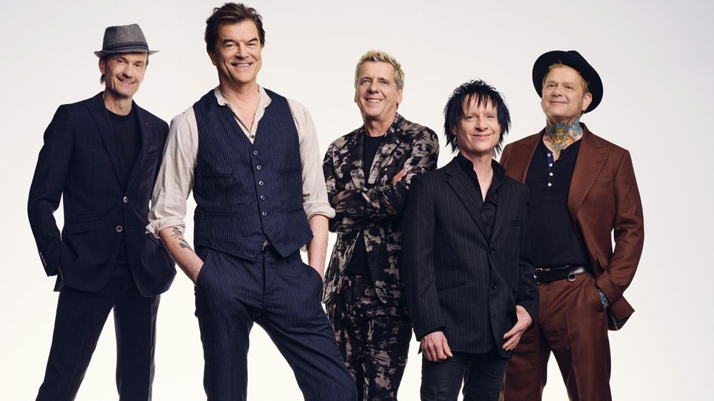 Vor einem hellen Hintergrund stehen die fünf Mitglieder der Band in Anzügen und lächeln. 