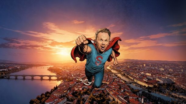 Ralf Schmitz fliegt als Superman über eine  Großstadt bei Sonnenuntergang. Es hat comichafte Züge. 