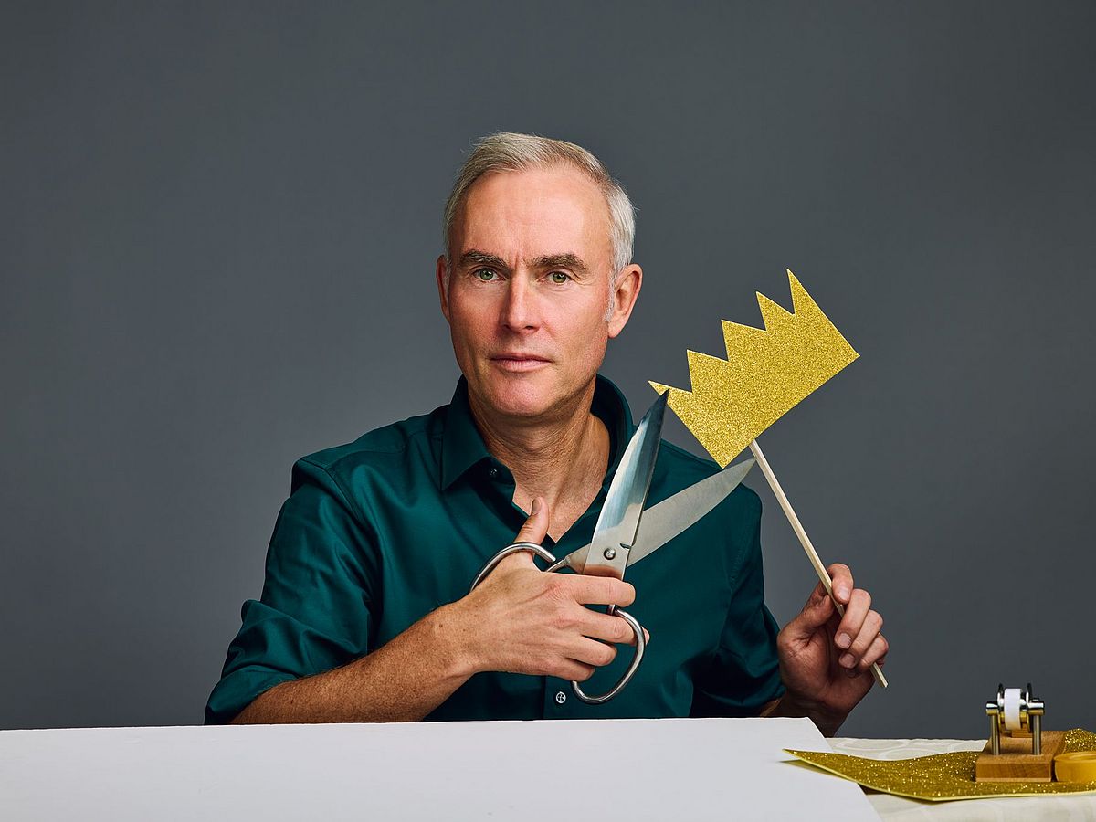 Johann König sitzt an einem Tisch vor einer grauen Wand. Er trägt ein dunkelgrünes Hemd. Bastelmaterial für die goldene Krone liegen auf dem Tisch, er hat eine fertige Krone in der Hand. Zudem noch eine anormal große Bastelschere. 