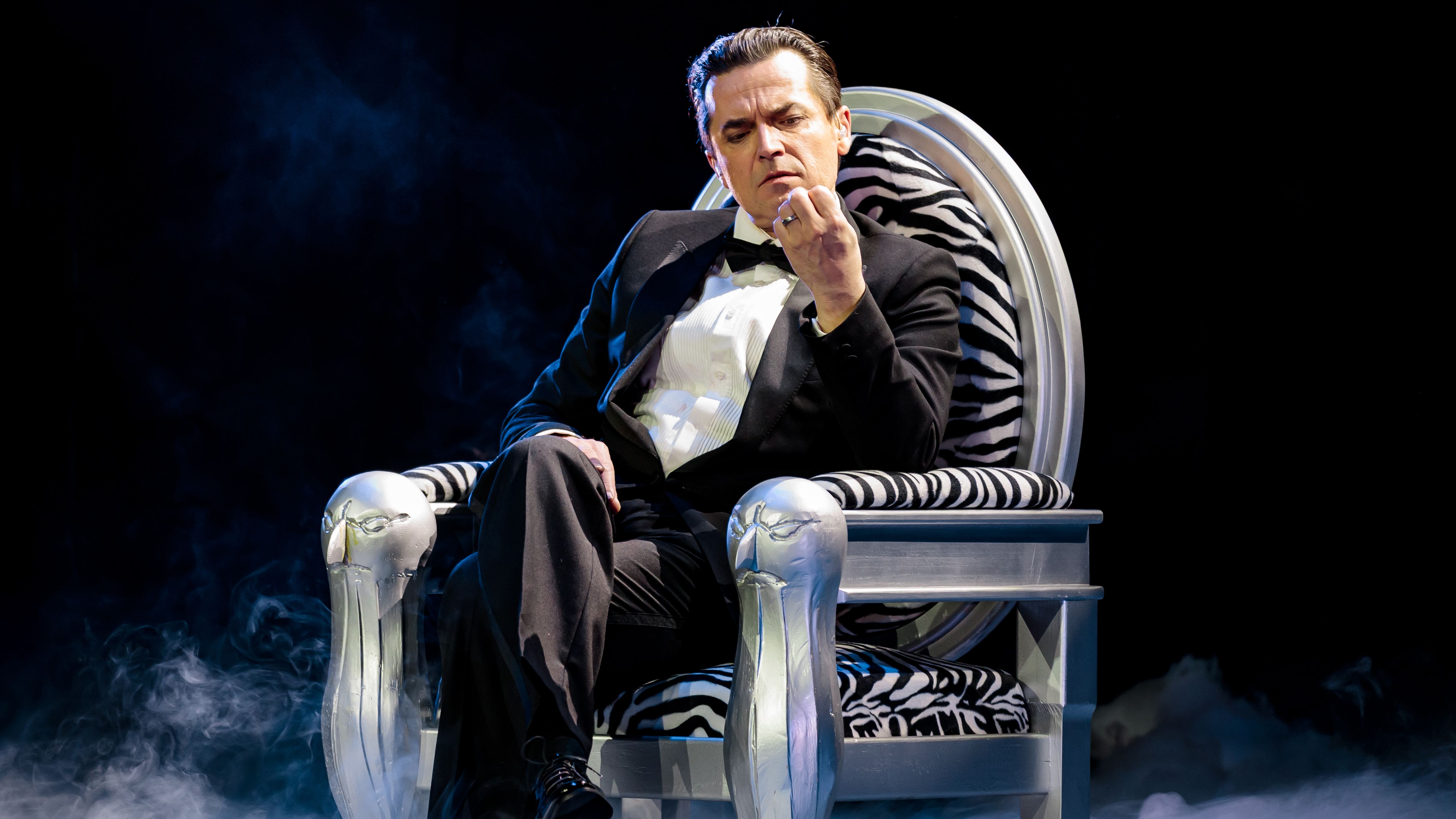 Falco sitzt auf einem thron-ähnlichen Stuhl, auf dem Boden ist Nebel. Der Falco-Darsteller trägt einen Anzug mit Fliege. 