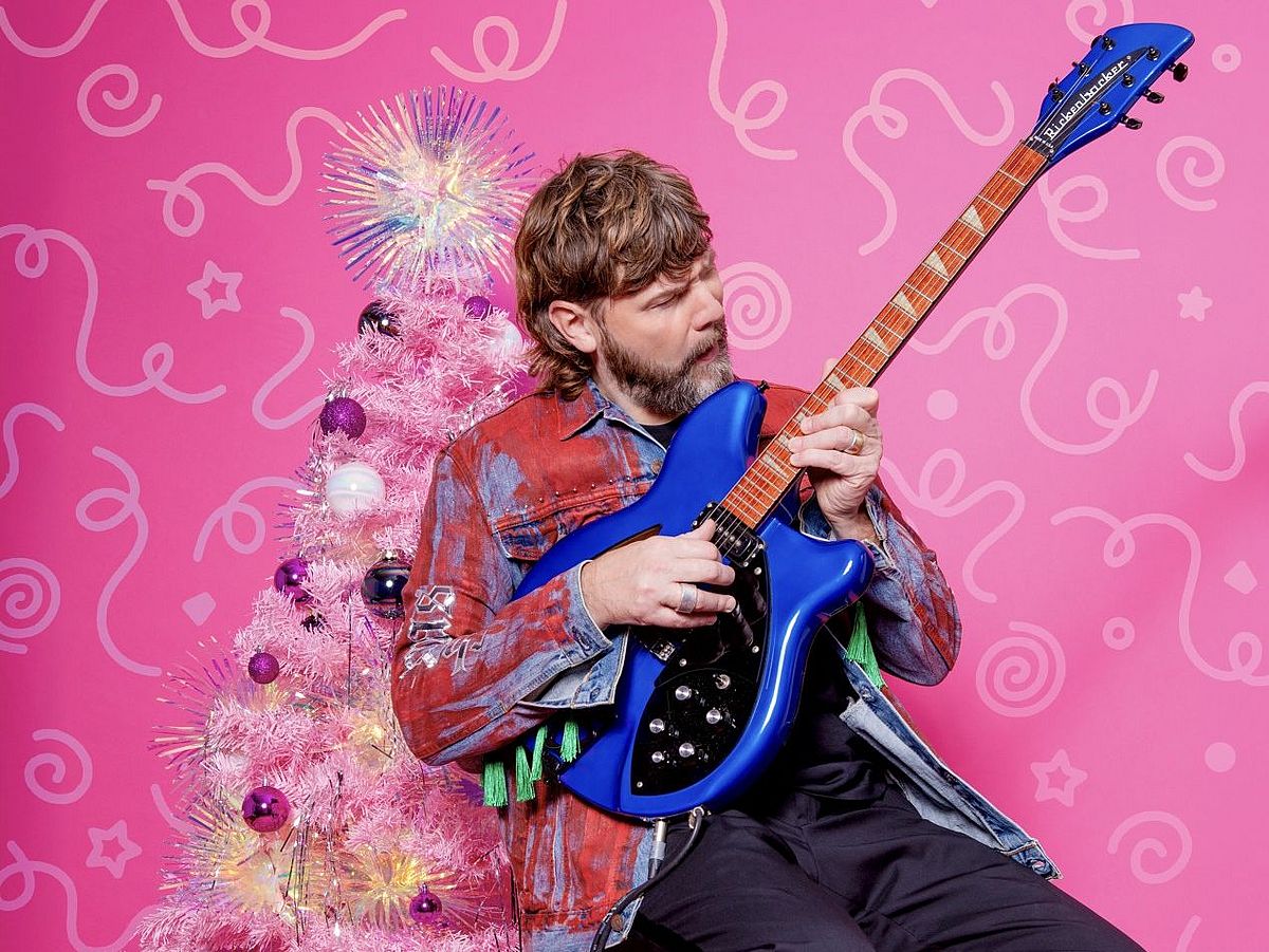 Rea Garvey sitzt auf einem Hocker mit einer E-Gitarre in der Hand. Hinter ihm ist ein künstlicher, weißer Weihnachtsbaum, der geschmückt ist. Der Hintergrund ist pink. 