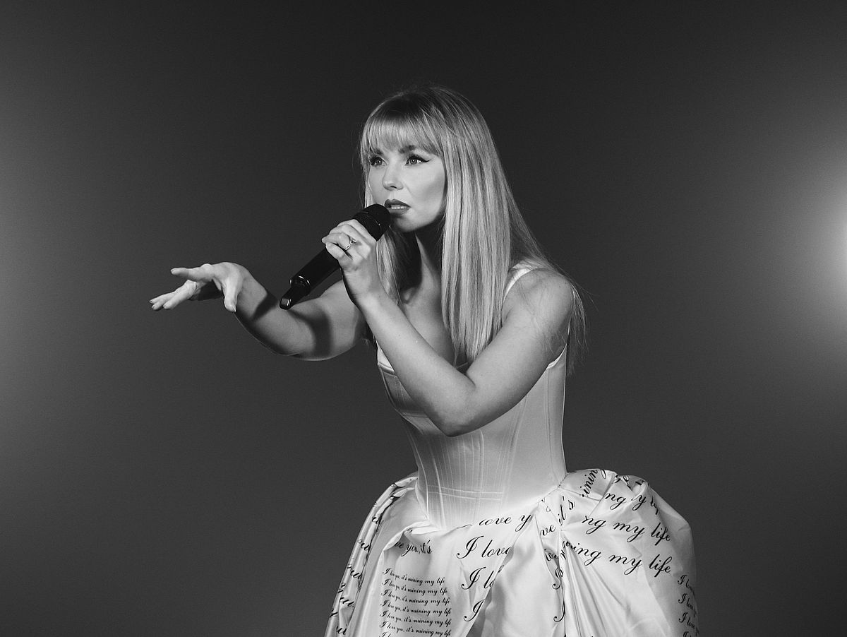 Schwarz-weißes Pressebild der Sängerin der Show. Sie trägt ein Satin-Kleid mit Stiefeletten und einer dunklen Strumpfhose. 