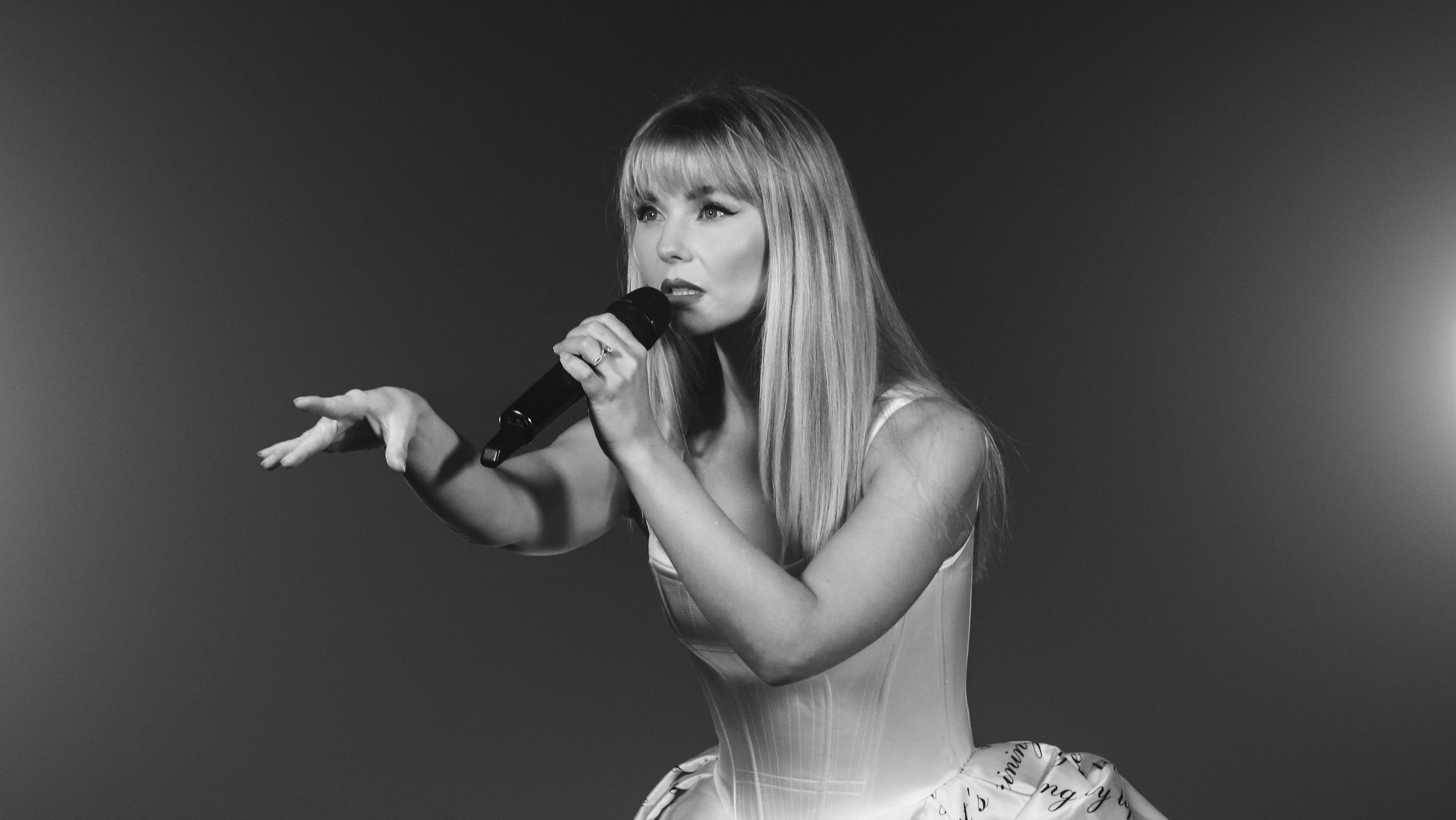 Schwarz-weißes Pressebild der Sängerin der Show. Sie trägt ein Satin-Kleid mit Stiefeletten und einer dunklen Strumpfhose. 